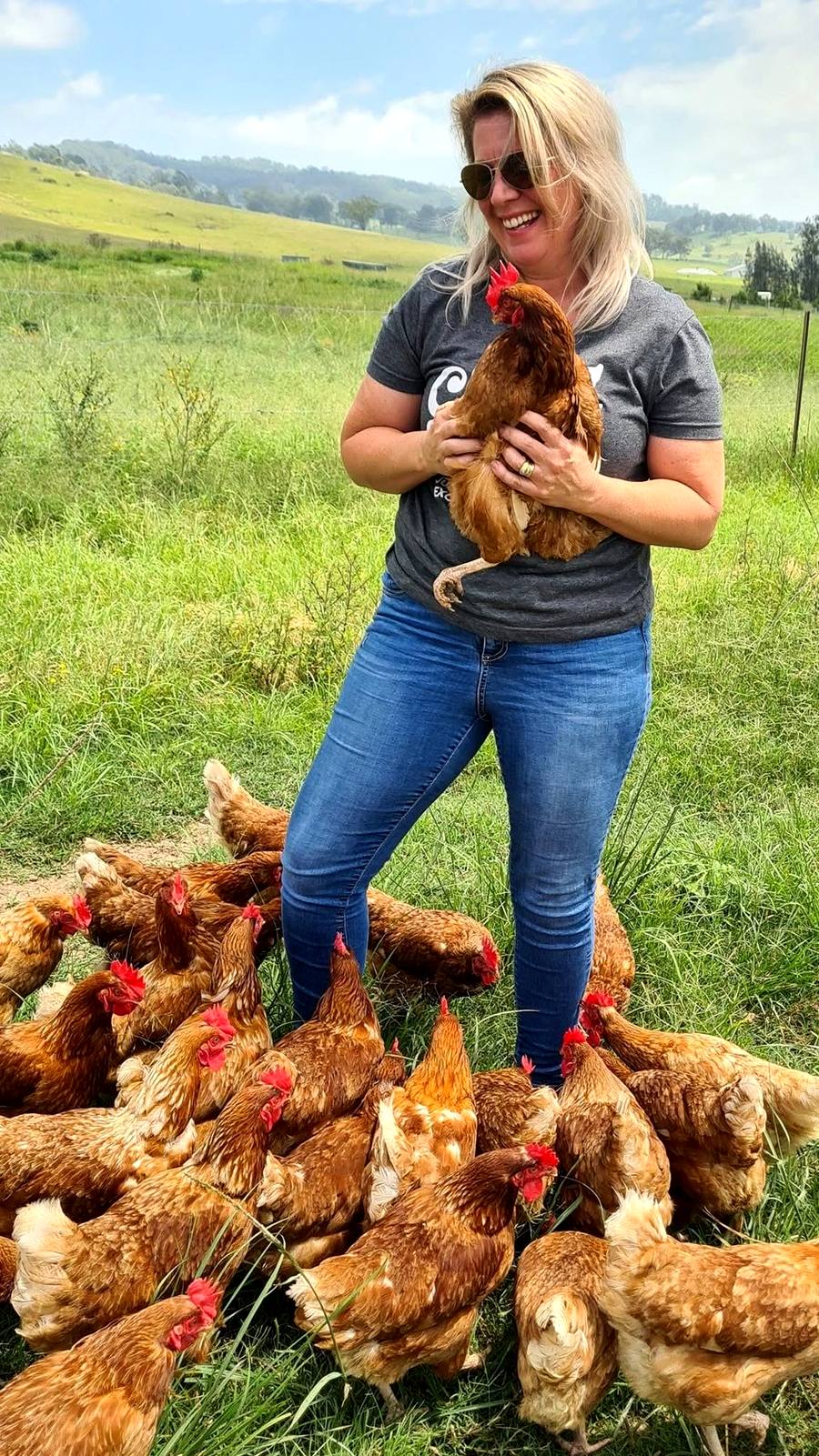 Simone standing in the paddock, laughing, a red hen in her arms and dozens more around her feet