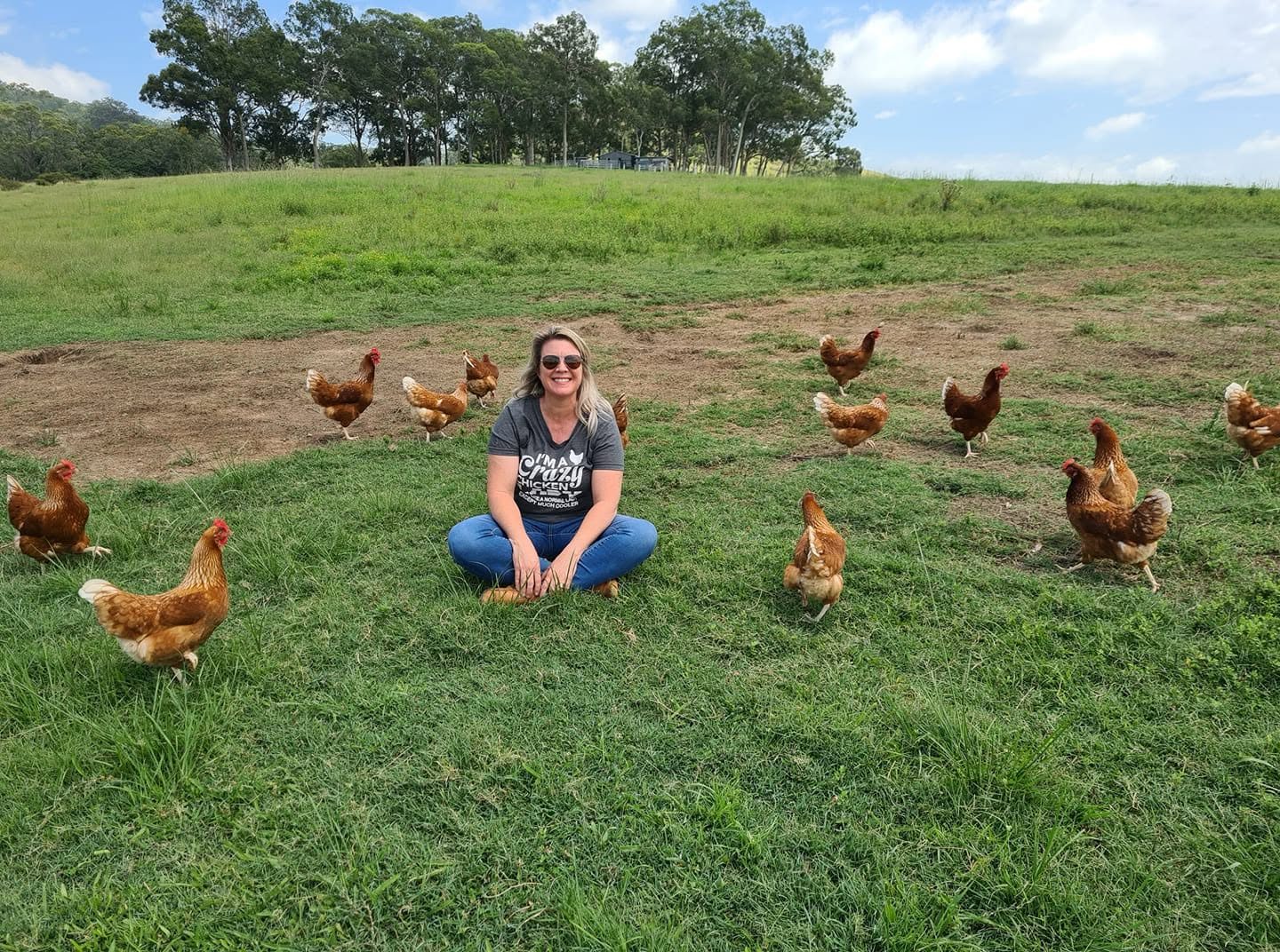 Simone sitting cross-legged in the grass, smiling, with her red hens wandering around her